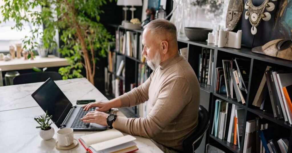 Middle-aged man working on a laptop at a modern home office desk with bookshelves, plants, and coffee nearby