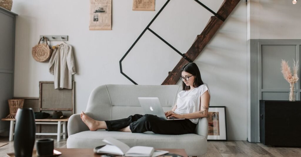 Woman using a laptop while relaxing on a sofa in a cozy modern living room with minimalist decor and stairs