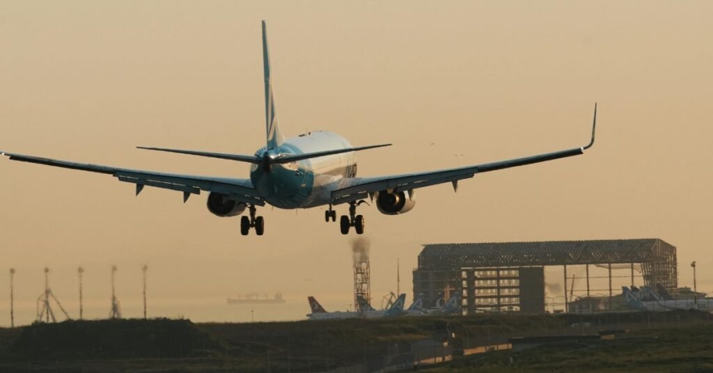 An airplane is landing at an airport during sunset, with warm sky tones and airport buildings visible in the background.